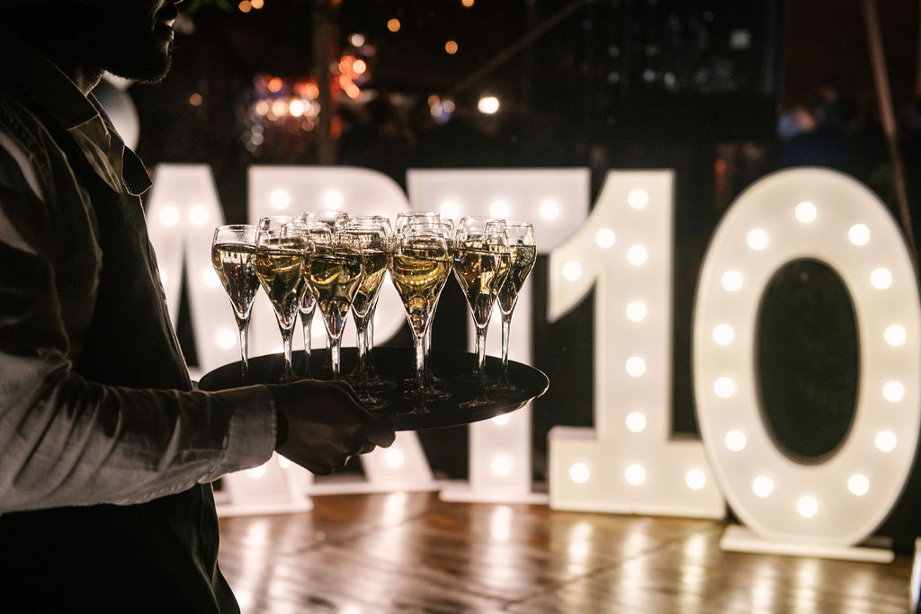 Serveur tenant un plateau de verres de champagne devant le logo d'Artone en lettres lumineuses géantes